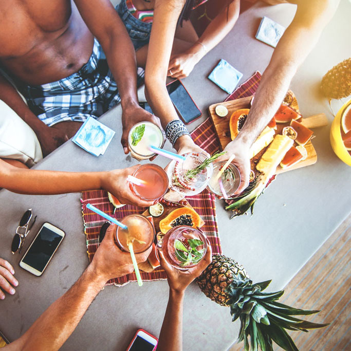 group of friends drinking  cocktails at a summer party sitting on a table toasting cheers, wearing casual beachwear. water melon pineapple and fruits on table. overhead view.