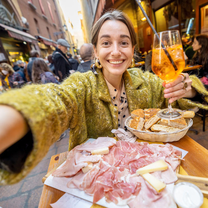 Happy woman taking selfie photo on phone while sitting with meat plate and cocktail at outdoor bar in Bologna city. Concept of Italian gastronomy and travel